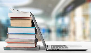 A stack of books sits next to an open laptop on a desk with a blurred background.