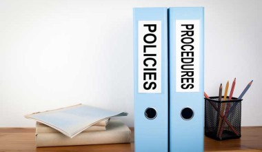 Two blue binders labeled Policies and Procedures sit on a desk next to folders and a cup holding pencils.