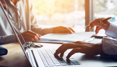 Two people sit at a desk with a laptop and documents, one person typing on the keyboard and the other holding a pen.