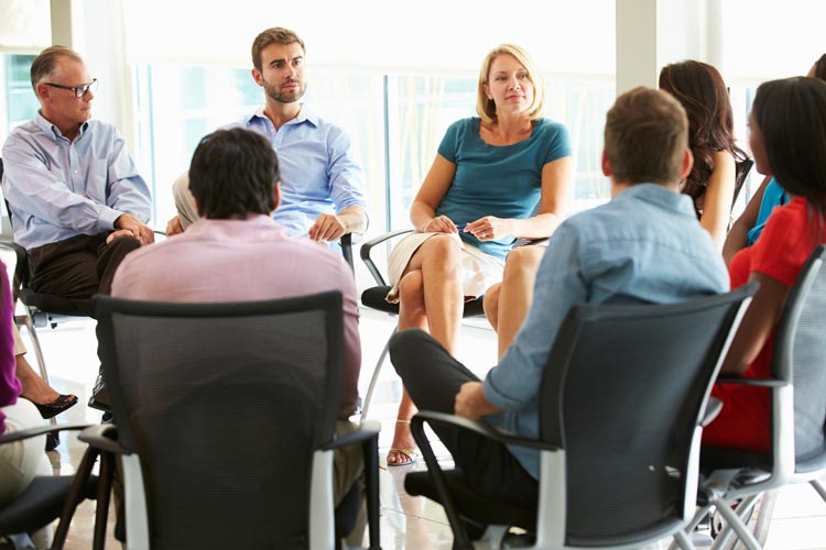A group of people sit in a circle having a discussion in a brightly lit office.