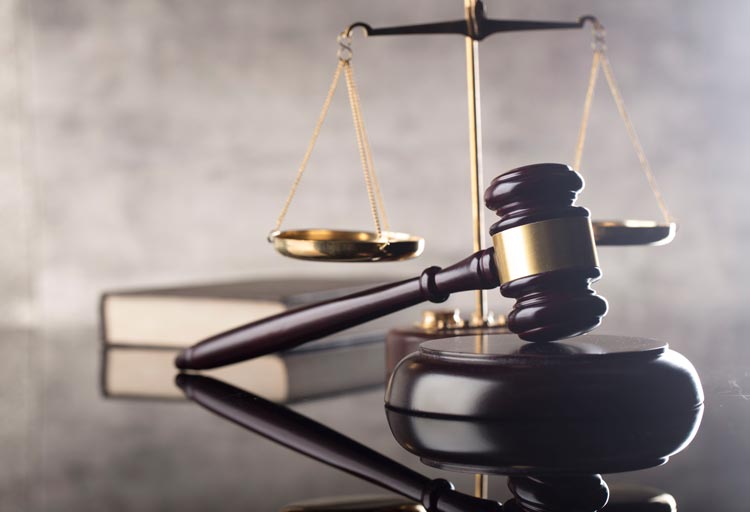 A judge’s gavel and block rest in front of balanced brass scales and stacked legal books on a glossy reflective surface.