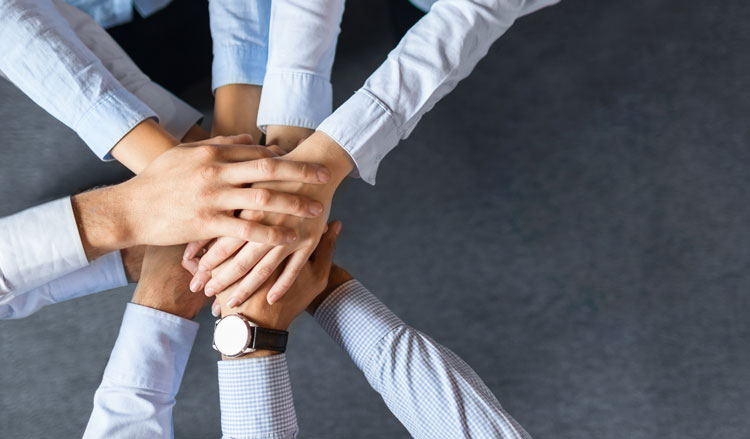 Five people stack their hands together in the center, viewed from above, representing teamwork and unity.