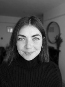 Woman with straight dark hair wearing a black turtleneck smiling in a softly lit room, photographed in black and white.