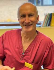Smiling man in pink scrubs with a name badge sits at a desk in front of a window and bookshelf.