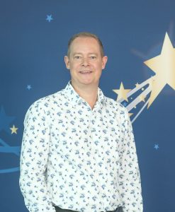 Smiling man wearing a white floral shirt stands in front of a blue background decorated with stars and shooting star graphics.