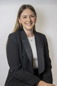 Smiling woman with long hair wearing a black blazer and white top sitting in front of a plain light background.