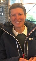 A smiling woman stands indoors, dressed in a navy jacket over a white collared shirt.