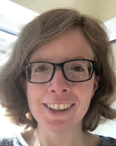 A person with short brown hair and glasses smiling indoors, illuminated by natural light.