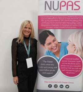 A woman smiles while standing beside a NUPAS banner displaying information on pregnancy advisory services and patient testimonials.