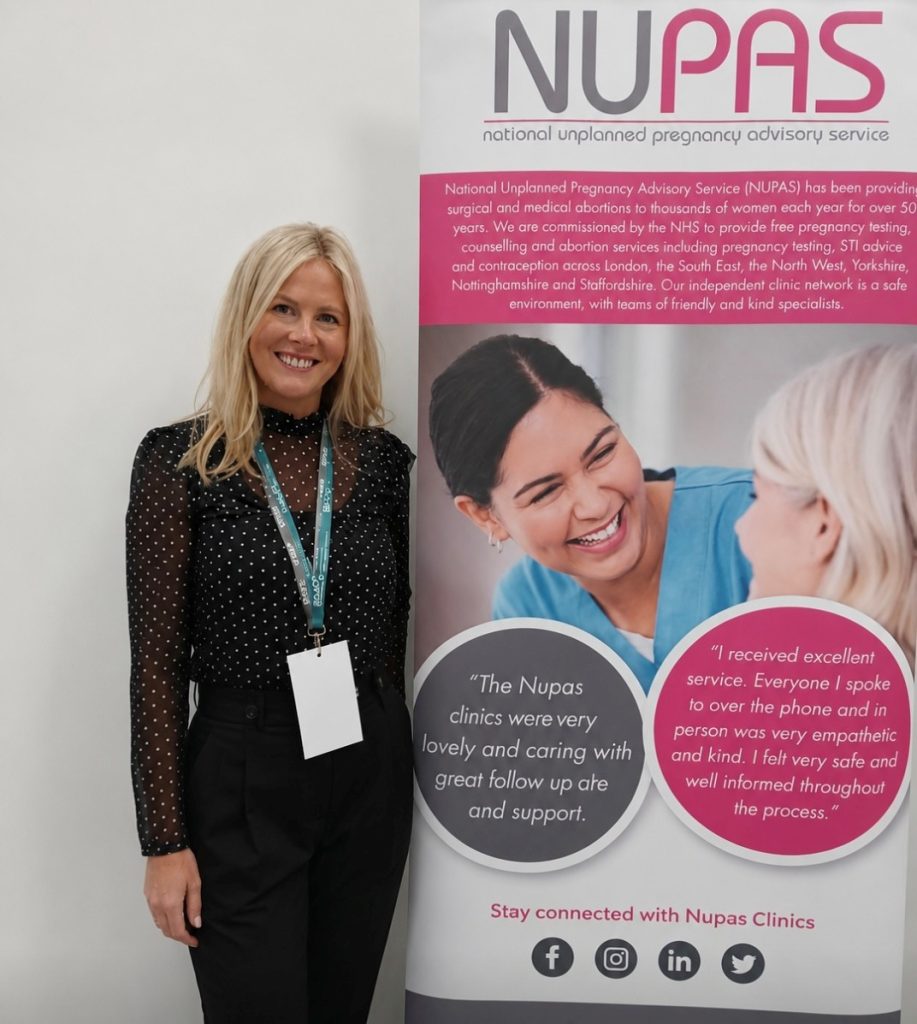 A woman smiles while standing beside a NUPAS banner displaying information on pregnancy advisory services and patient testimonials.