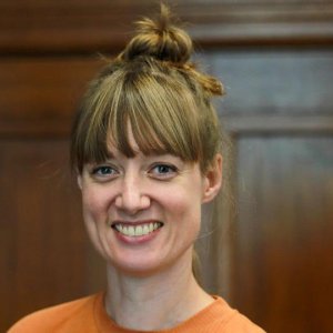 A woman with light brown hair in a topknot, smiling and wearing an orange shirt, stands in front of a wooden background.